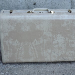 May include: A vintage beige suitcase with a handle and gold-colored latches. The suitcase has a textured surface with wear and tear marks. The handle and latches are made of metal. The suitcase is rectangular in shape.