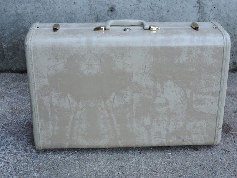 May include: A vintage beige suitcase with a handle and gold-colored latches. The suitcase has a textured surface with wear and tear marks. The handle and latches are made of metal. The suitcase is rectangular in shape.