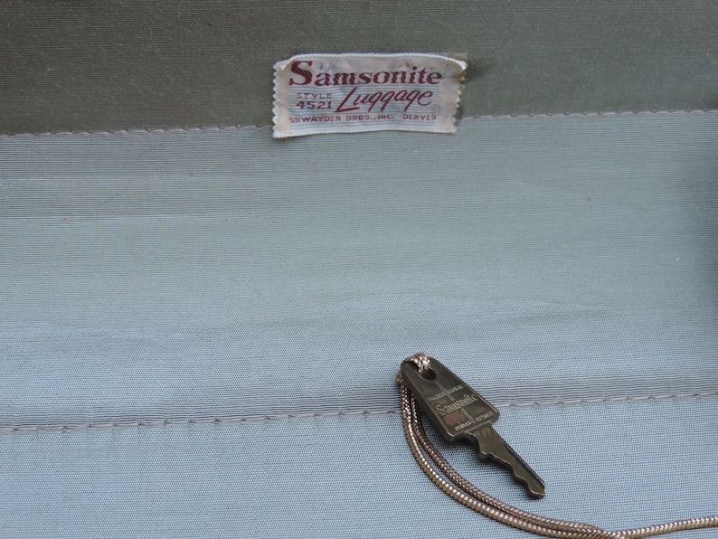 May include: Vintage Samsonite luggage interior with a key attached. The interior is a light blue-green color with a stitched seam. A tag reads "Samsonite Luggage, Style 4521, SWAYDER BROS. INC. DENVER."