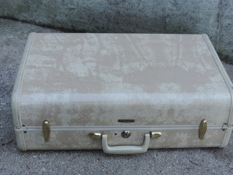 May include: Tan vintage suitcase with a textured pattern and gold-colored hardware. The suitcase has a handle and a lock. The corners are reinforced with white trim. A classic travel accessory.