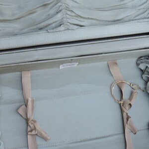 May include: Open vintage suitcase with a light green interior and a silver frame. The interior features a pleated fabric lining, beige ribbons, and a gold-colored circular object. The suitcase is open, revealing its contents.