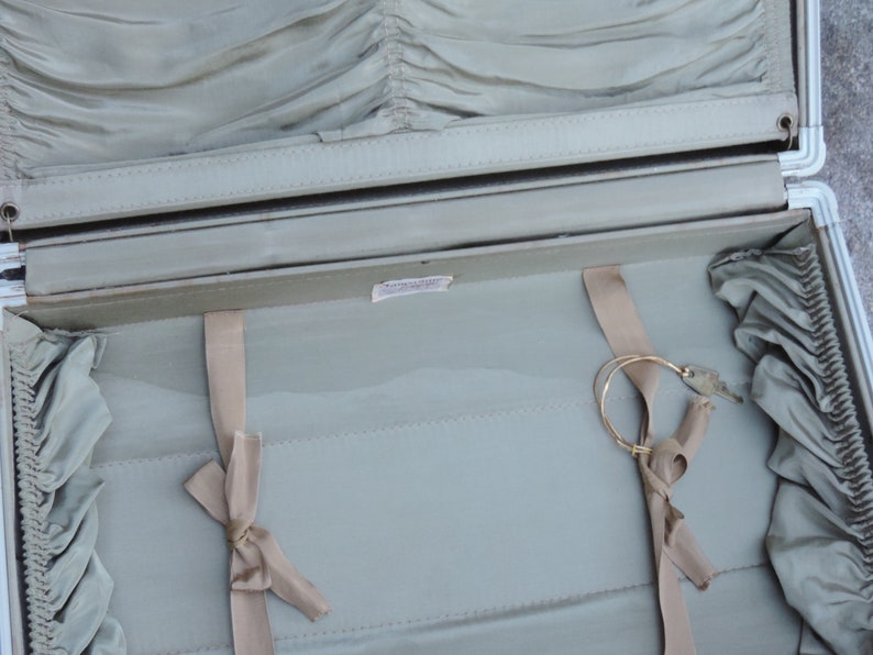 May include: Open vintage suitcase with a light green interior and a silver frame. The interior features a pleated fabric lining, beige ribbons, and a gold-colored circular object. The suitcase is open, revealing its contents.
