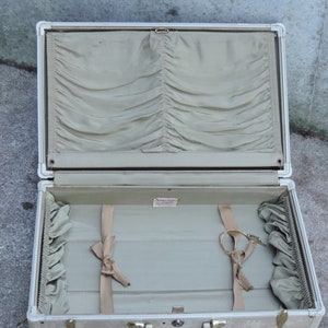 May include: Open vintage suitcase with a light beige exterior and silver trim. The interior is lined with a light green fabric, featuring a pleated top section and ribbon tie-downs. Includes a key and a small label.