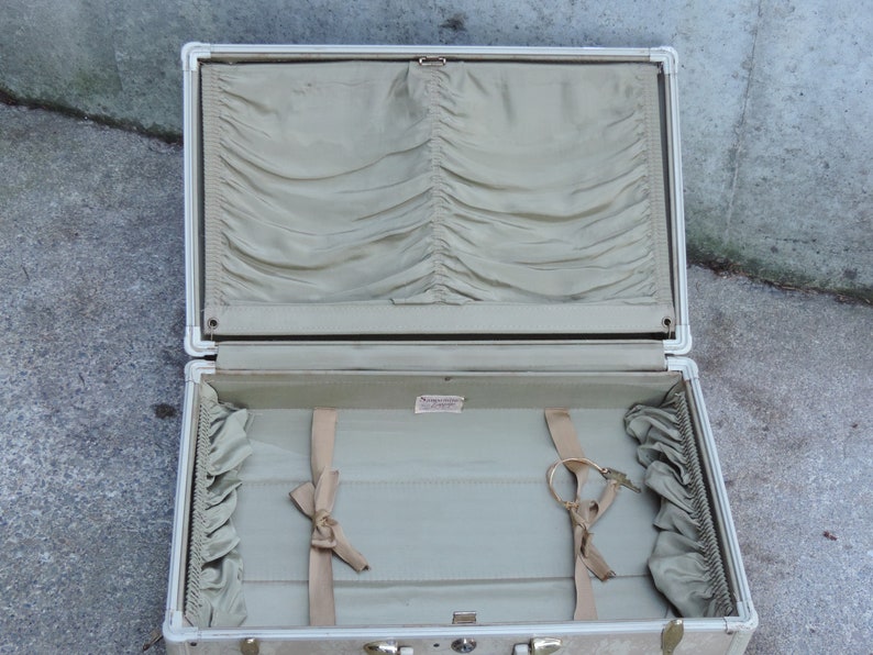 May include: Open vintage suitcase with a light beige exterior and silver trim. The interior is lined with a light green fabric, featuring a pleated top section and ribbon tie-downs. Includes a key and a small label.