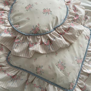 May include: A round pillow and a rectangular pillow featuring a cream background with a pink rose and blue ribbon floral pattern. Both pillows have a ruffled edge with a light pink striped design and are trimmed with a thin blue piping border.