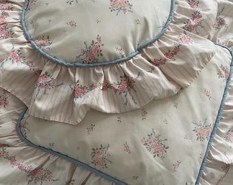 May include: A round pillow and a rectangular pillow featuring a cream background with a pink rose and blue ribbon floral pattern. Both pillows have a ruffled edge with a light pink striped design and are trimmed with a thin blue piping border.