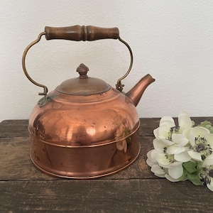 May include: A polished copper tea kettle featuring a rounded body, a short spout, and a removable lid with a small knob. The kettle has a curved brass handle with a dark brown wooden grip centered at the top for lifting and pouring.