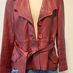 May include: A burgundy leather jacket with a belted waist and red buttons. The jacket has a classic collar and two front pockets. A baseball cap rests on the mannequin's shoulder. The jacket is paired with blue denim jeans.