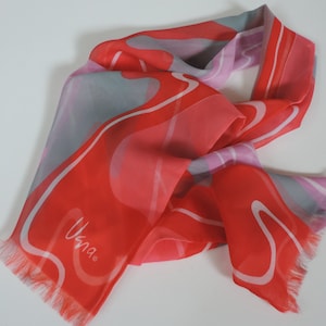 May include: A sheer scarf in red, pink, and grey, with white wavy lines. The scarf has a fringed edge and the word "Usna" in white script. A fashionable accessory.