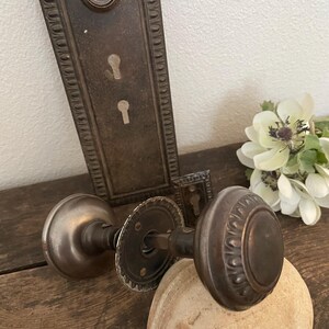 May include: An antique bronze door knob set featuring a round knob with a decorative scalloped edge, a matching rectangular backplate with two keyholes, and a smaller rectangular escutcheon plate, all displaying a weathered, dark metallic finish.