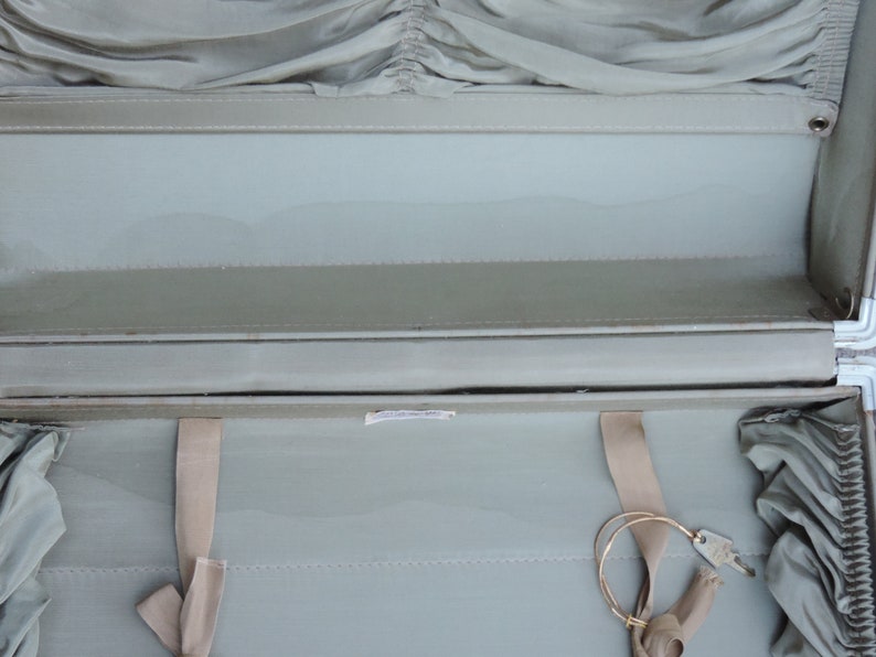 May include: Open vintage suitcase with a light green interior and exterior. The suitcase has a metal frame and hinges. Beige fabric straps and a key are visible inside. The interior has a pleated fabric detail.