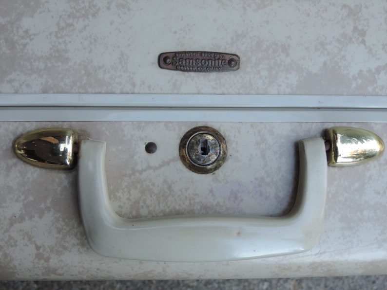 May include: Close-up of a vintage Samsonite suitcase. The suitcase has a cream-colored exterior with a speckled pattern. It features a brass lock, handle, and accents. The Samsonite logo is visible.