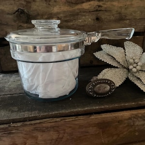 May include: A clear glass saucepan with a matching glass lid and a long glass handle. The pot features a silver-toned metal band around the upper rim. The interior of the glass pot is filled with white paper, and a decorative metal oval piece sits nearby.