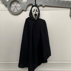 May include: A hanging Ghostface figure with a white mask featuring a black mouth and eye openings, wearing a black hooded robe. Behind the figure is a large, gray, decorative key-shaped wall hanging with the text Boo... You Rang? printed on it.