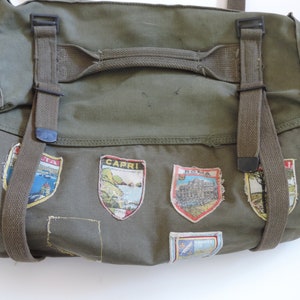 May include: Olive green canvas duffel bag with shoulder straps and decorative patches. The patches feature colorful illustrations of various locations. The bag has a top handle and adjustable straps with black buckles.