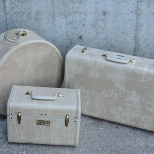 May include: Three vintage beige suitcases of varying sizes. The set includes a round hatbox-style case, a rectangular suitcase, and a smaller rectangular case. All cases have gold-toned hardware and handles.
