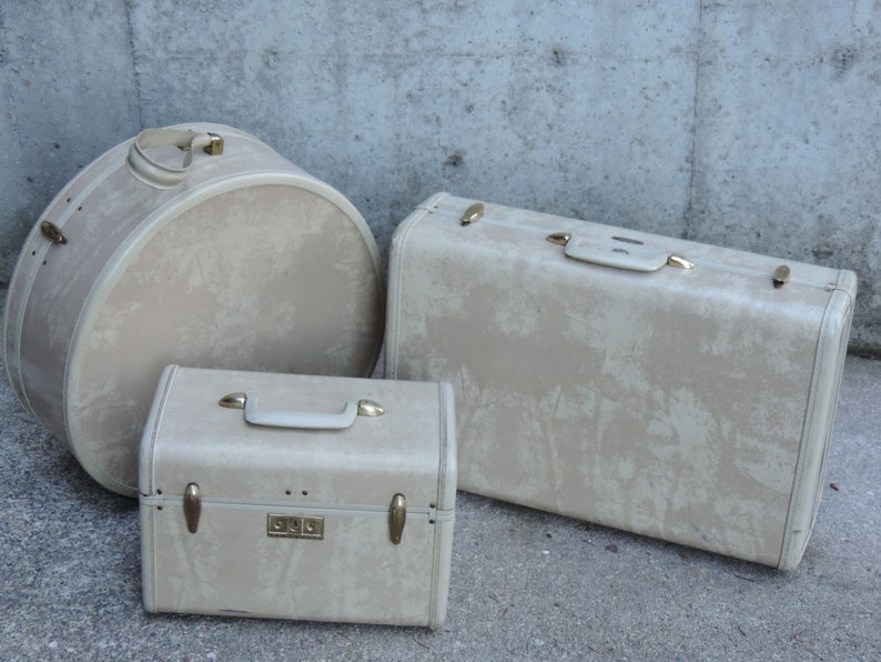 May include: Three vintage beige suitcases of varying sizes. The set includes a round hatbox-style case, a rectangular suitcase, and a smaller rectangular case. All cases have gold-toned hardware and handles.