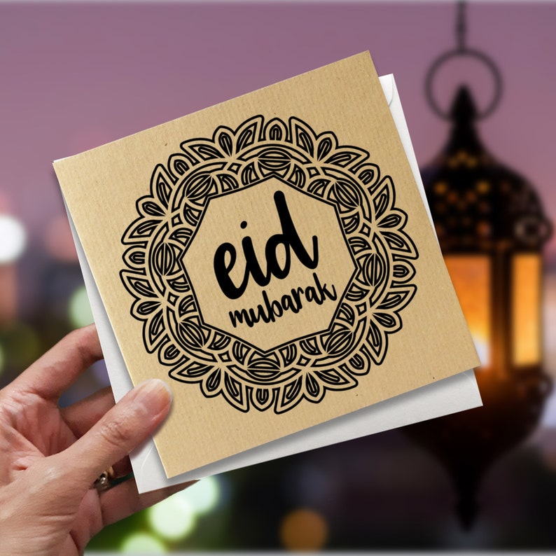 May include: A square greeting card with the words "eid mubarak" in black script, surrounded by a decorative black mandala design. The card is held in a hand, with a blurred background featuring a lantern.