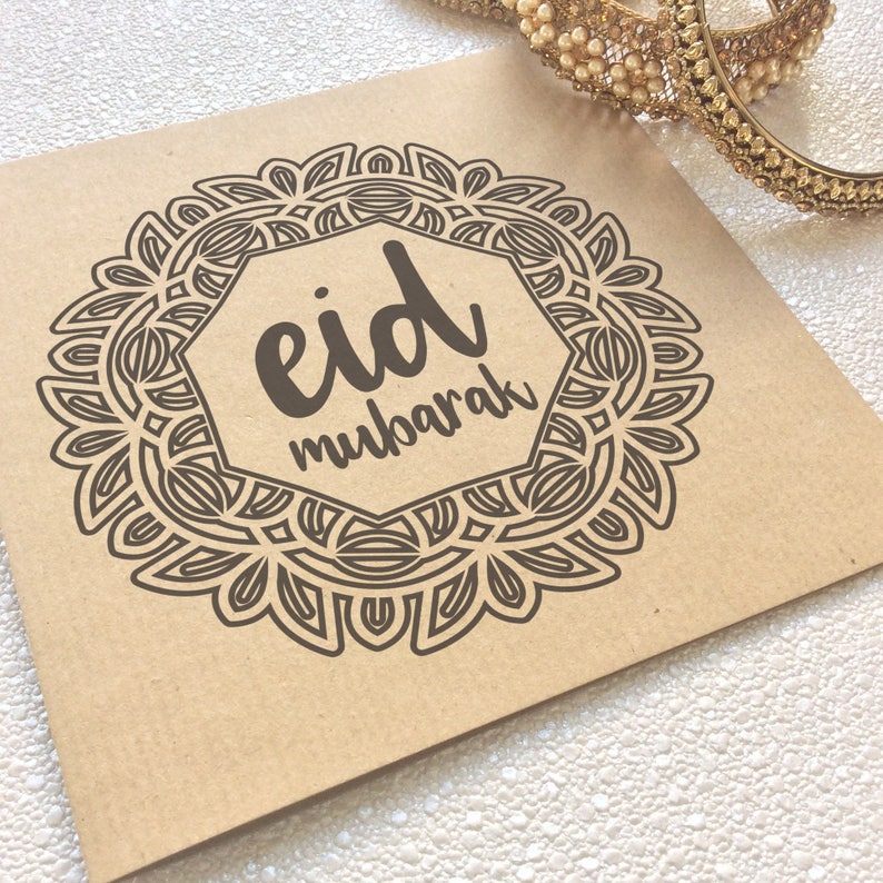 May include: A square, tan card with a brown, ornate circular design and the words "Eid Mubarak" in a stylized font. Gold bangles with pearl accents are in the background. The card is likely for the Eid holiday.