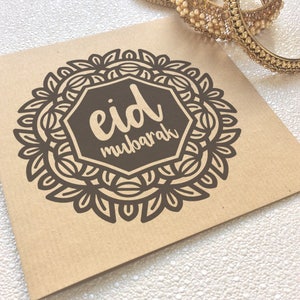 May include: A brown card with a black and white mandala design. The mandala has a hexagon in the center with the text "eid mubarak" in a white cursive font.