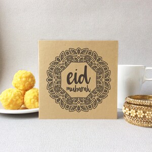May include: A square, kraft paper card with the words "Eid Mubarak" in a decorative frame. The image also includes a white mug, gold bangles, and a plate of yellow sweets.