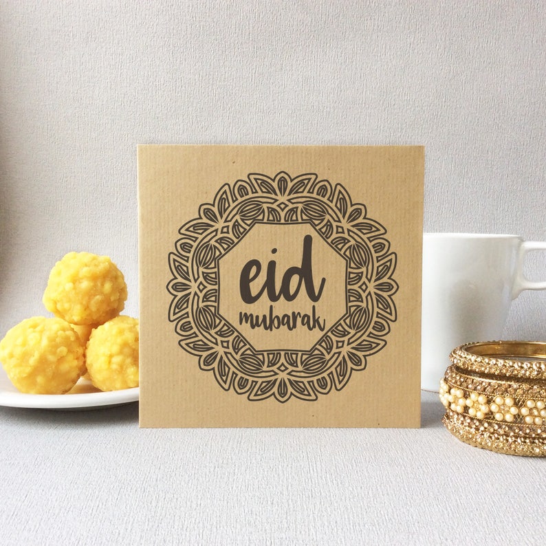 May include: A square, kraft paper card with the words "Eid Mubarak" in a decorative frame. The image also includes a white mug, gold bangles, and a plate of yellow sweets.