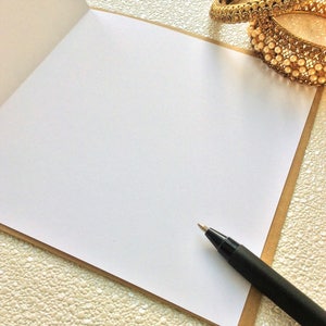 May include: A blank white card with a brown border, a black pen, and a gold and white beaded bracelet on a textured white surface.