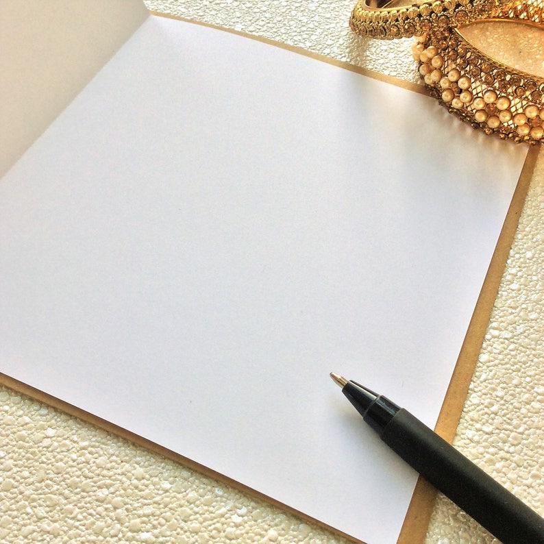 May include: A blank white card with a brown border, a black pen, and a gold and white beaded bracelet on a textured white surface.