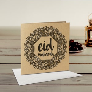 May include: A square greeting card with the words "Eid Mubarak" in black script, surrounded by a decorative circular border. The card is made of brown cardstock and rests on a white envelope. A lit lantern and dates are in the background.