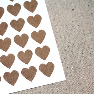 May include: A collection of small, brown paper heart cutouts arranged on a white surface. The hearts are evenly spaced and have a natural, rustic appearance. The background is a textured, neutral-toned fabric.