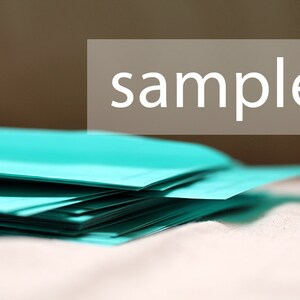 May include: A stack of teal-colored paper sheets. The word "sample" is displayed in white text on a rectangular, light brown background. The paper is stacked on a white surface.