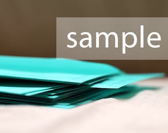 May include: A stack of teal-colored paper sheets. The word "sample" is displayed in white text on a rectangular, light brown background. The paper is stacked on a white surface.