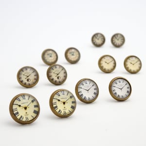 May include: A collection of antique-style clock face cabinet knobs with brass-colored metal rims. The clock faces feature Roman numerals and various designs, including a moon phase. The knobs are arranged on a white background.