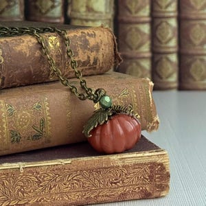 May include: A bronze-toned necklace featuring a small, orange pumpkin pendant. The pumpkin has a detailed, textured surface and is accented with a green bead and a bronze leaf. The necklace rests on antique books.