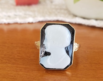 May include: A gold ring with a black and white cameo featuring a profile of a person. The cameo is set in a rectangular frame.