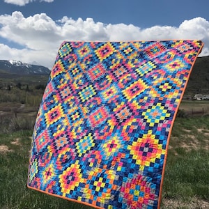 May include: A colorful quilt with a geometric pattern in shades of blue, yellow, pink, and orange. The quilt is made of squares and rectangles and has a unique, swirling design. The quilt is displayed outdoors against a backdrop of green grass and a blue sky with white clouds.