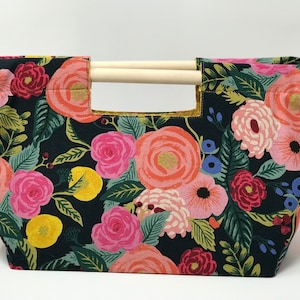 Op de afbeelding: Een zwarte stoffen tas met een bloemenprint met roze, rode, gele en groene bloemen. De tas heeft houten handvatten.