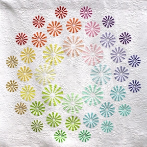 Op de afbeelding: Een wit quilt met een cirkelvormig ontwerp van kleurrijke windmolens. De windmolens zijn in een spiraalvormig patroon gerangschikt, beginnend met rode en oranje windmolens in het midden en overgaand naar paarse en blauwe windmolens aan de buitenrand.