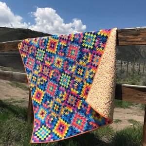 May include: A colorful quilt with a geometric pattern in shades of blue, yellow, orange, pink, and gray. The quilt is hanging on a wooden fence against a blue sky with white clouds.