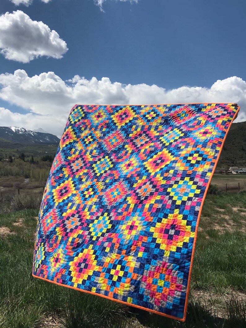 May include: A colorful quilt with a geometric pattern in shades of blue, yellow, pink, and orange. The quilt is made of squares and rectangles and has a unique, swirling design. The quilt is displayed outdoors against a backdrop of green grass and a blue sky with white clouds.