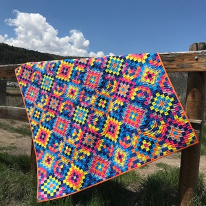 May include: A colourful quilt with a geometric pattern in shades of blue, yellow, pink, orange, and grey. The quilt is hanging on a wooden fence against a blue sky with white clouds.