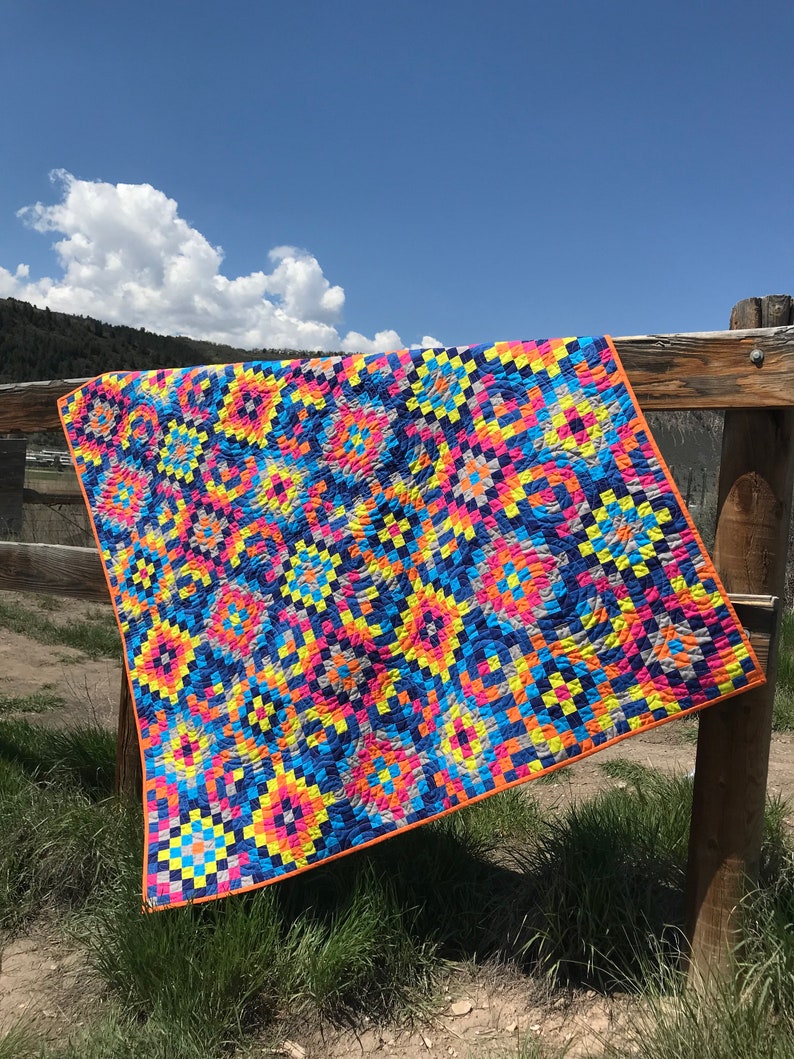 May include: A colorful quilt with a geometric pattern in shades of blue, yellow, pink, orange, and gray. The quilt is hanging on a wooden fence against a blue sky with white clouds.