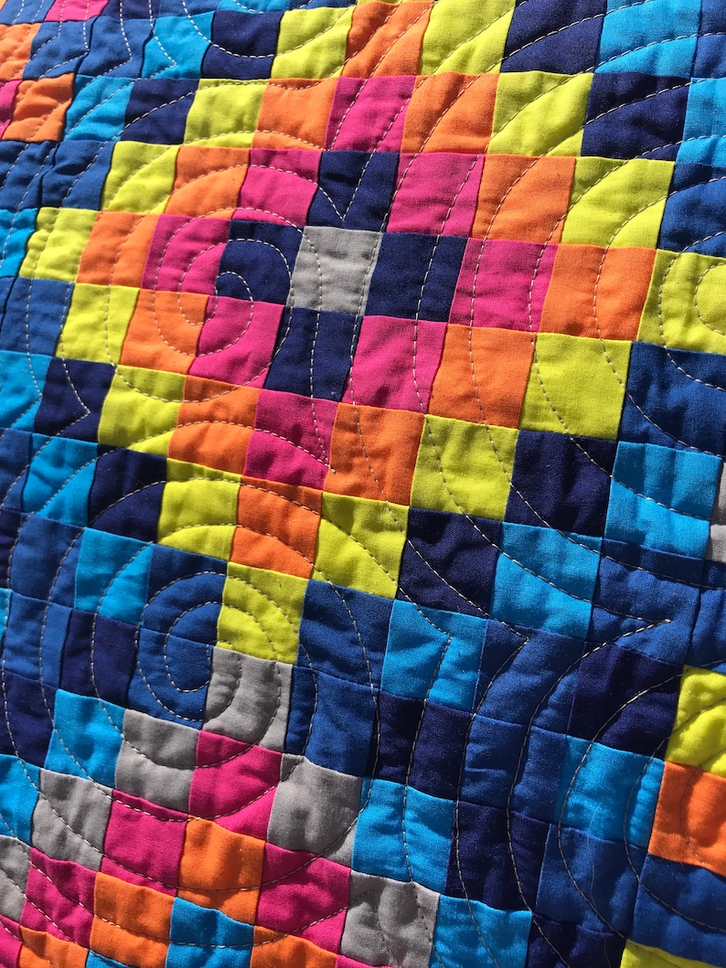 May include: A colorful quilt with a patchwork design featuring squares of blue, green, orange, pink, and gray fabric. The quilt is quilted with a circular pattern.