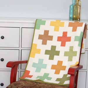 Pode incluir: Um edredom branco com um design de patchwork que apresenta sinais de mais coloridos em tons de verde, laranja, amarelo e marrom. O edredom está drapeado sobre uma cadeira vermelha com um almofada estampada.