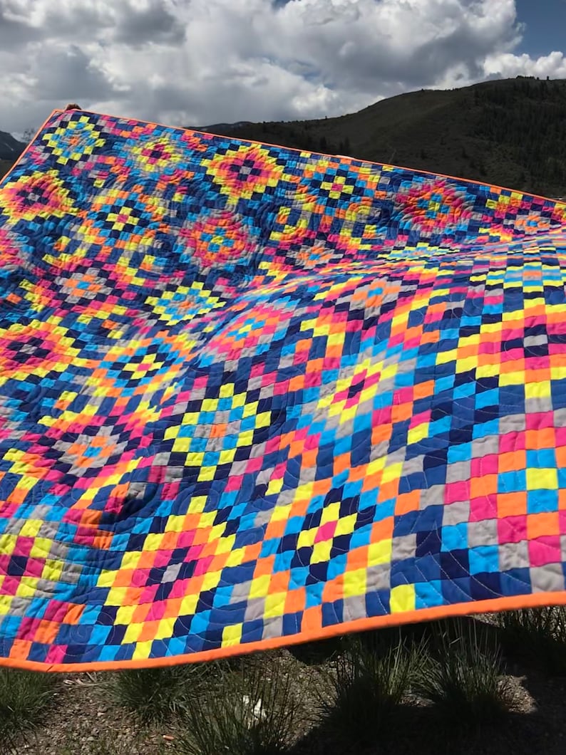 May include: A colorful quilt with a geometric pattern of squares and rectangles in shades of blue, yellow, pink, orange, and gray. The quilt is folded and draped over a grassy area.