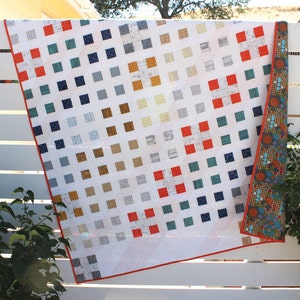 Può includere: Un quilt bianco con un motivo geometrico di quadrati in vari colori, tra cui rosso, blu, verde e marrone. Il quilt è appeso a una recinzione bianca.