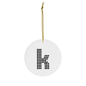 Könnte beinhalten: Weißer Keramik-Anhänger mit einem schwarzen Buchstaben K aus kleinen Kreisen. Der Anhänger hat eine goldene Schnur zum Aufhängen.