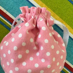 May include: A pink fabric drawstring bag with white polka dots. The bag has a white drawstring and is made of a soft, cotton fabric.