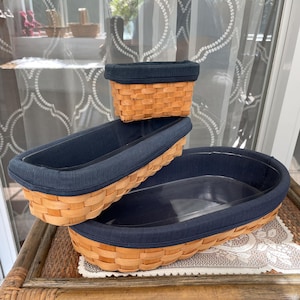 May include: Three woven wicker baskets with navy blue fabric lining. The largest basket is oval, the middle one is rectangular, and the smallest is square. The baskets are displayed on a bamboo table with a decorative lace doily.