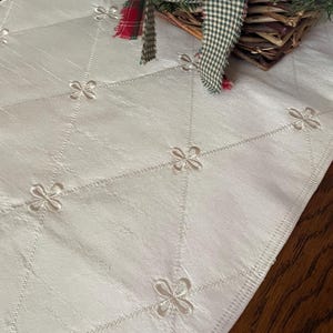 May include: Ivory-colored table runner with embroidered floral accents. The runner features a diamond pattern with delicate flower-shaped embroidery at each intersection. A decorative basket with greenery and plaid ribbon sits on the runner.